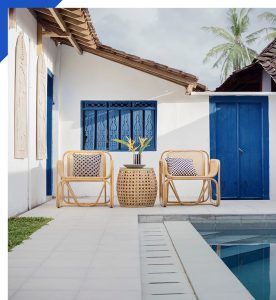 Two woven chairs and a small table beside a blue door and pool in a bright courtyard