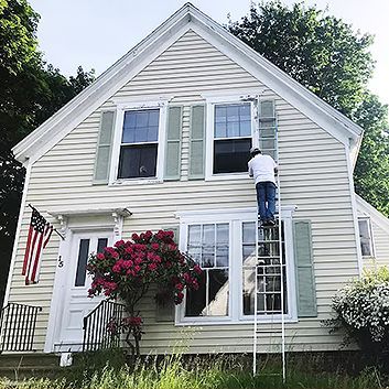 Person on a ladder painting the upper siding of a white house with green shutters