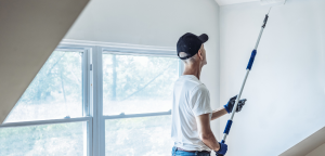 Person painting a white wall near a bright window using a roller brush