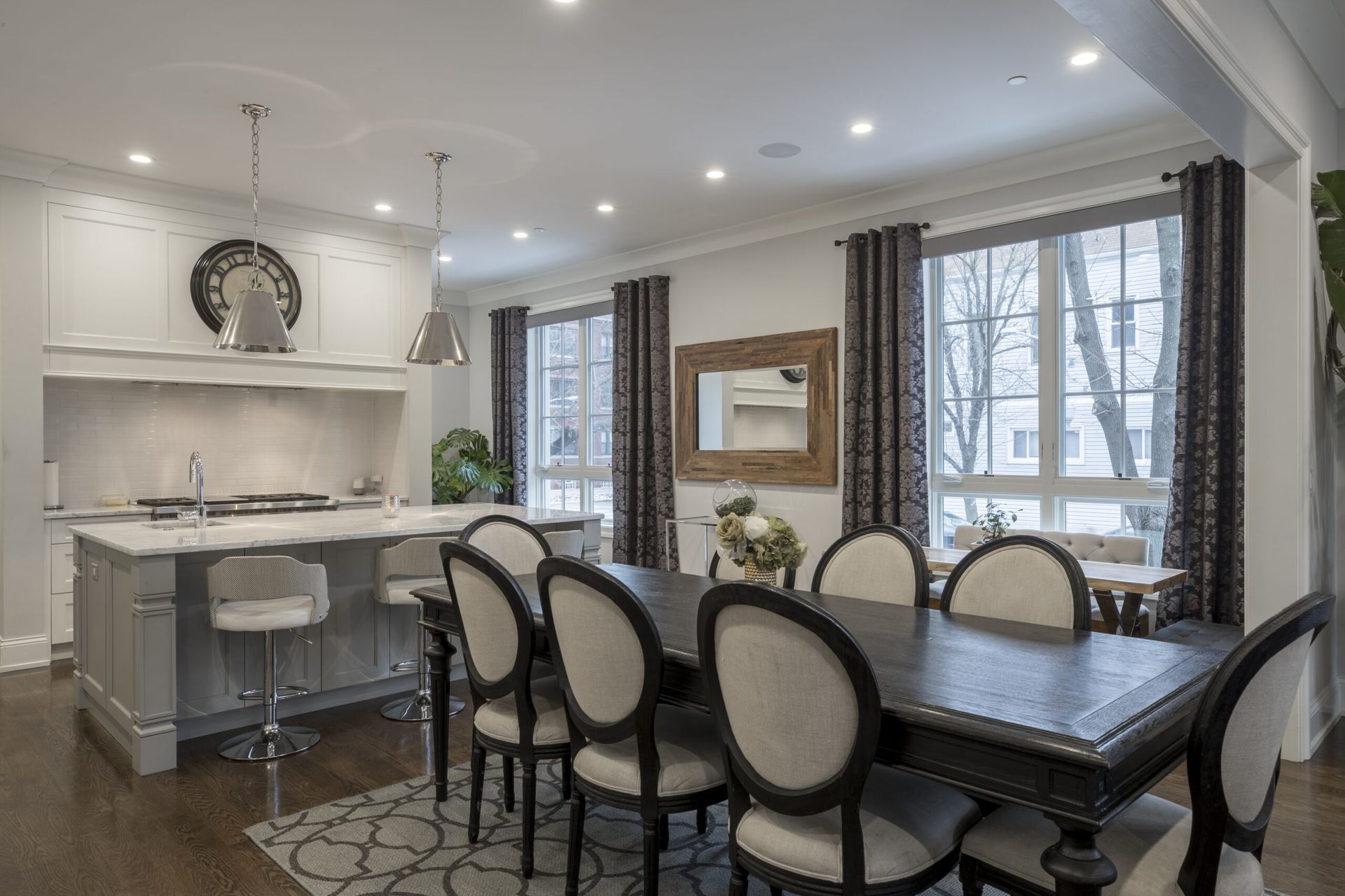 Bright dining room with a dark table, cream chairs, kitchen island, and large windows with gray drapes.