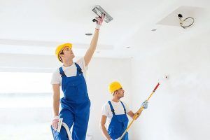 Two painters in blue overalls and yellow hard hats painting a white ceiling indoors