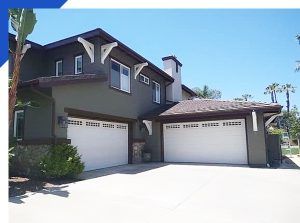 Two-story suburban house with dark trim, white garage doors, and a bright driveway under a blue sky