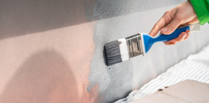 Hand holding a blue paintbrush, applying white paint to a wall edge