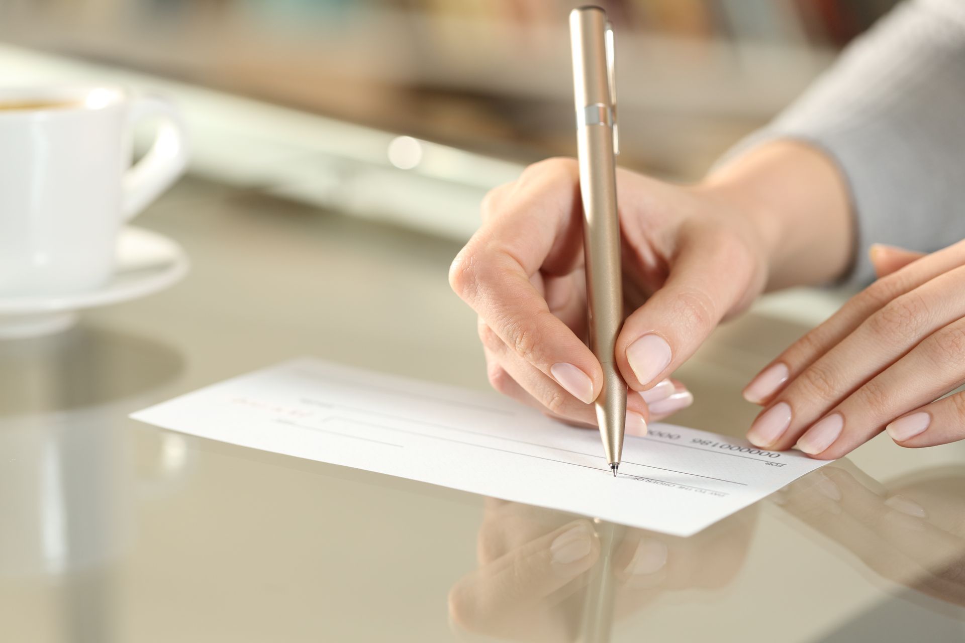 Person's hand writing with a pen on a check, with a coffee mug in the background.