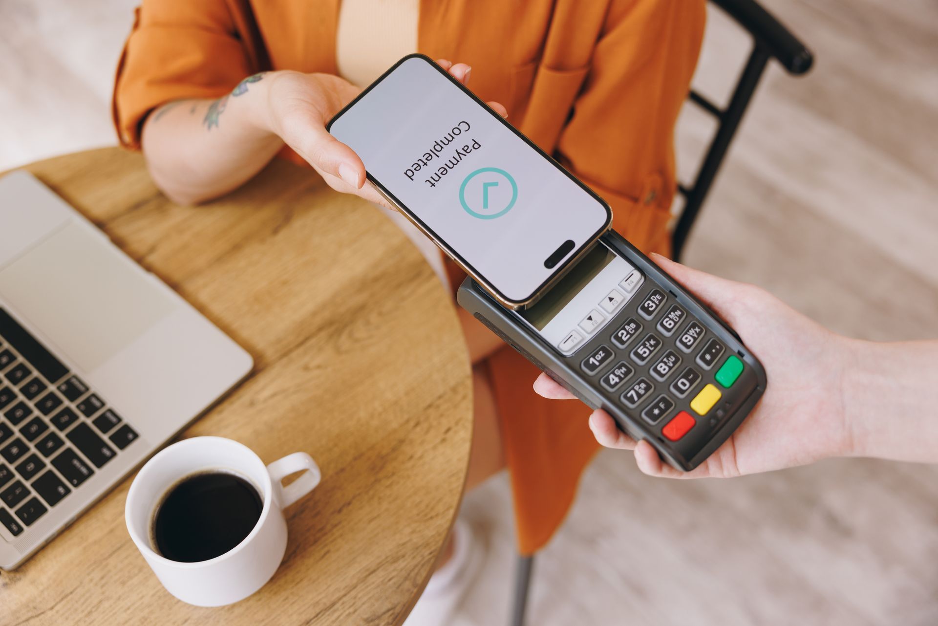 Person paying with phone at a cafe; phone over payment terminal, coffee and laptop nearby.