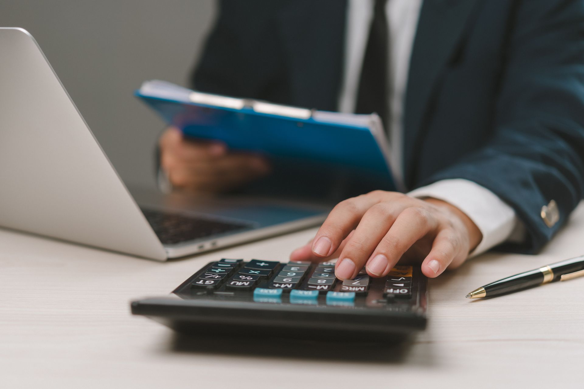 Person in suit using a calculator near a laptop and pen, holding a blue file.