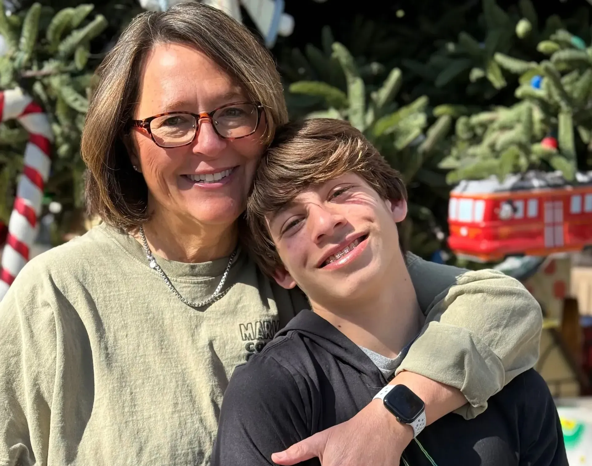 A smiling person with glasses embraces a younger person in front of a holiday-themed tree with a toy trolley ornament.