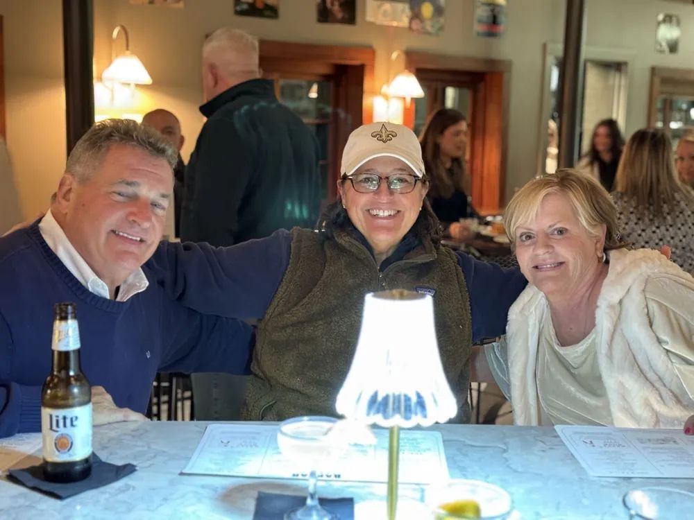 Three friends smiling at a restaurant table with a bottle of beer and a glowing lamp