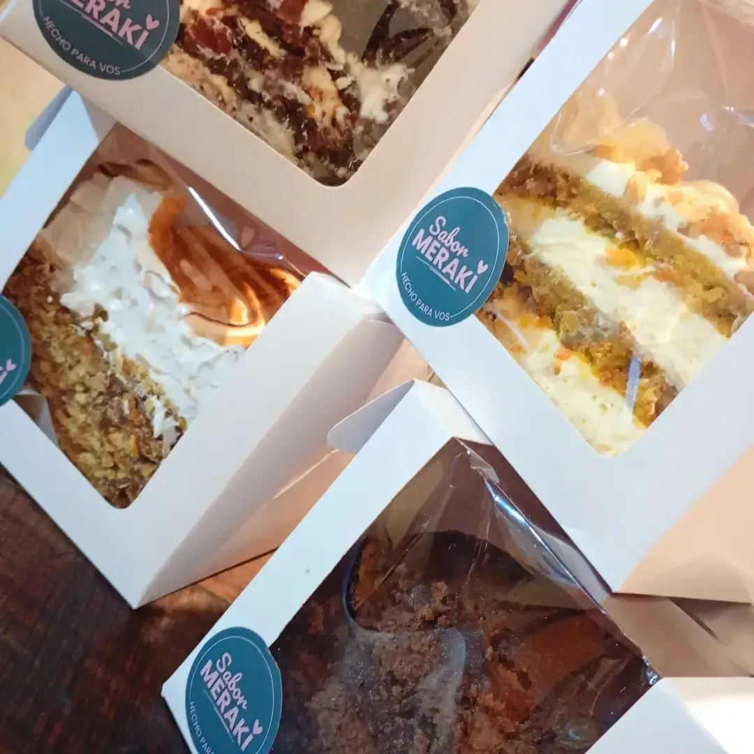 Porciones variadas de pastel en cajas, con glaseado blanco y etiquetas azules, sobre una mesa.