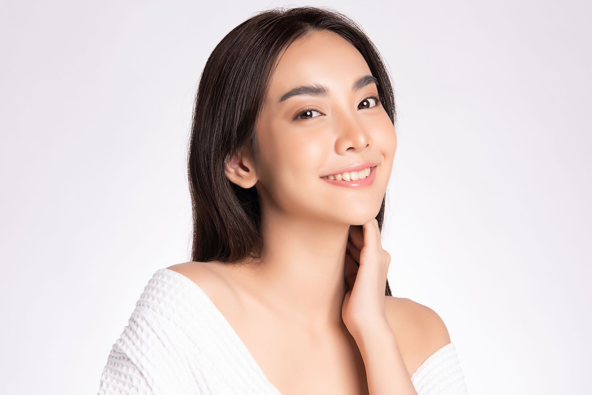 Smiling woman with long dark hair touching her neck, wearing a white off-shoulder top against a light background