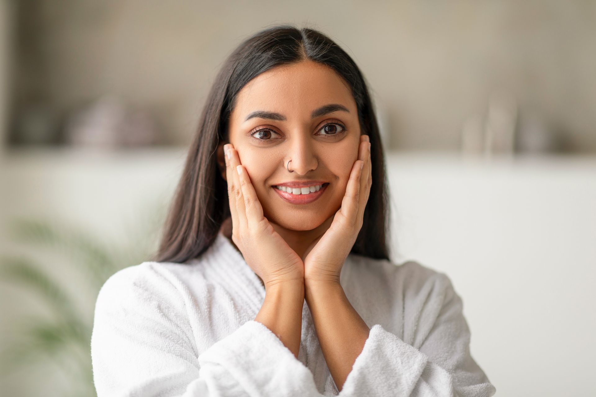 A woman in a bathrobe is smiling and touching her face.