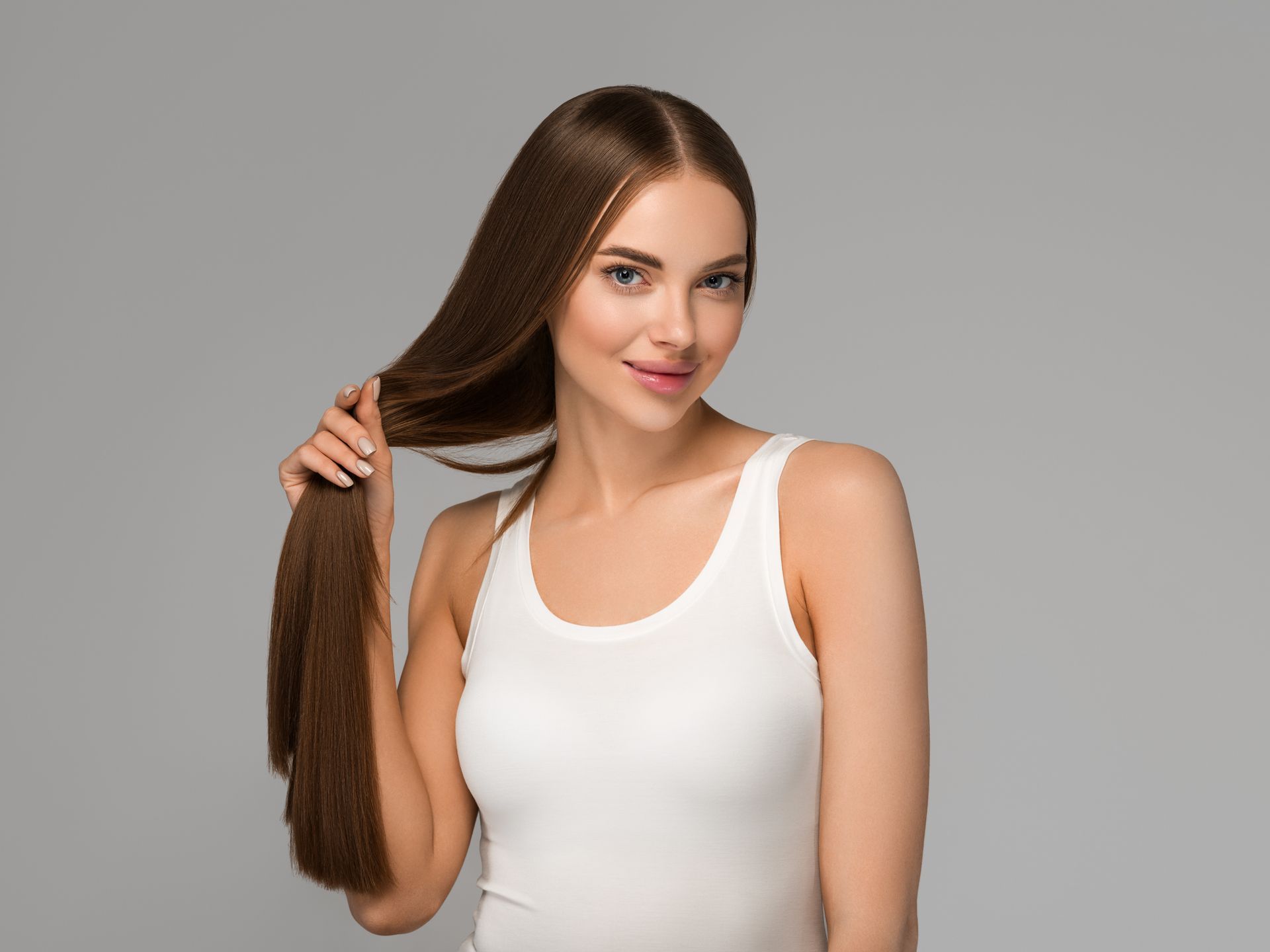 A woman in a white tank top is holding her long hair.