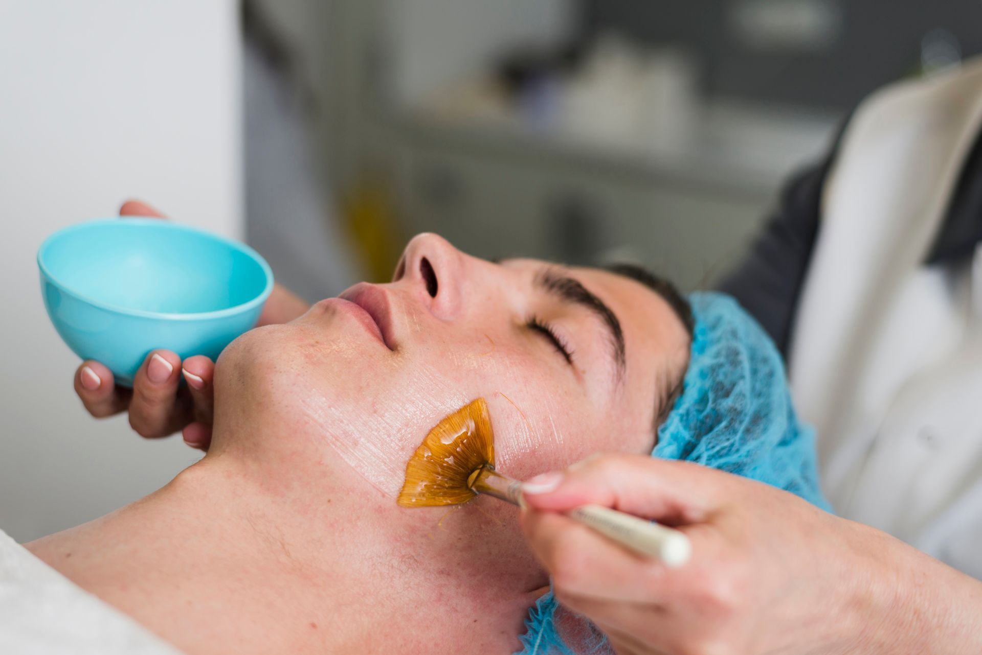 Facial treatment with a brush applying golden mask to a reclining person holding a blue bowl