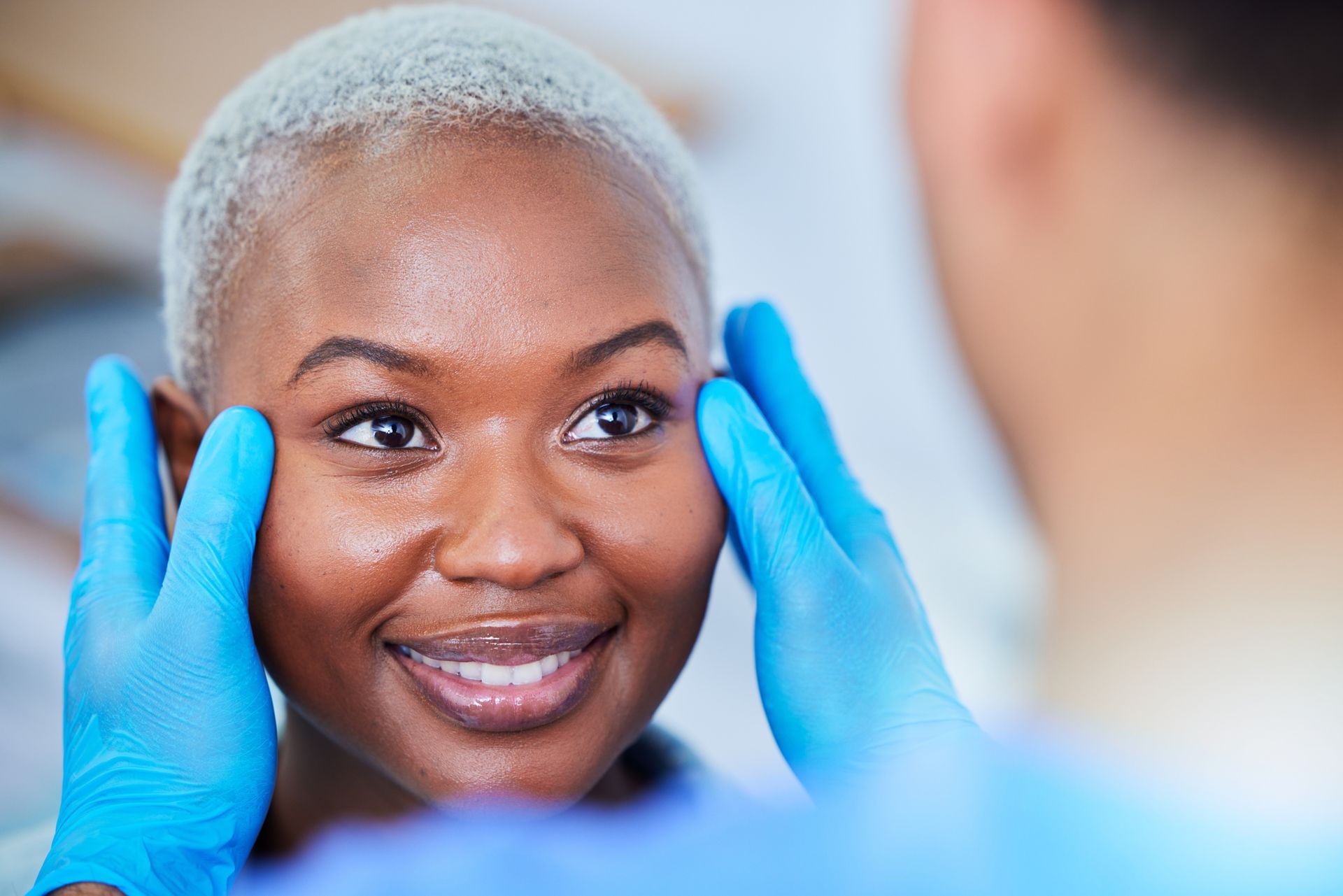 Blue-gloved hands gently holding a smiling person's face in a bright clinical setting