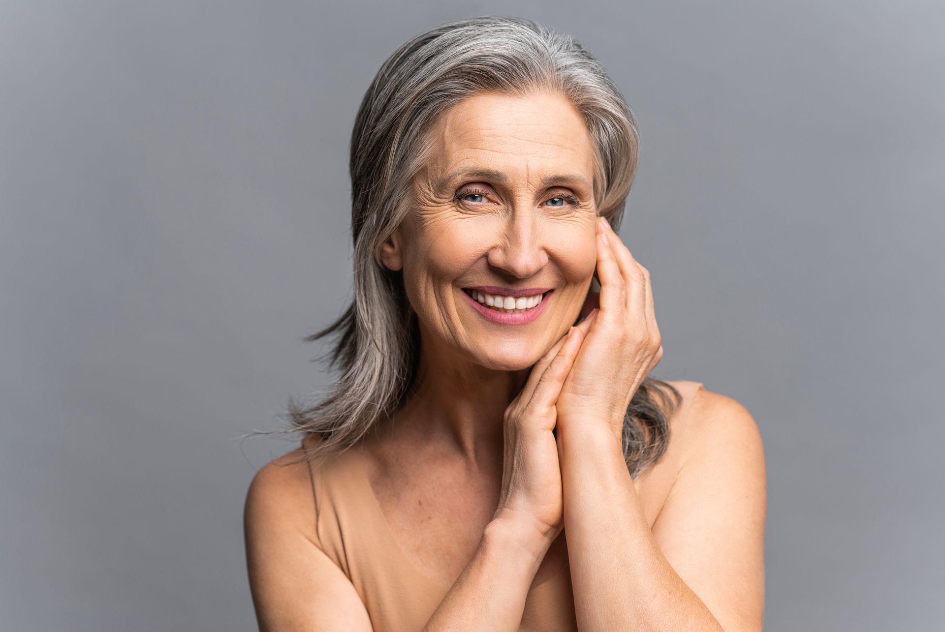 A smiling person with grey hair holds their hands against their cheek against a plain grey background.