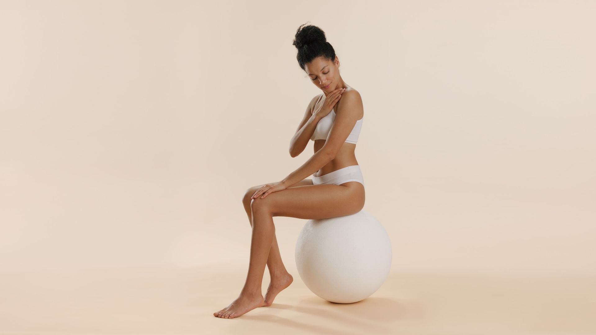 A person with a bun hairstyle sits on a white exercise ball against a beige background, looking down at their shoulder.