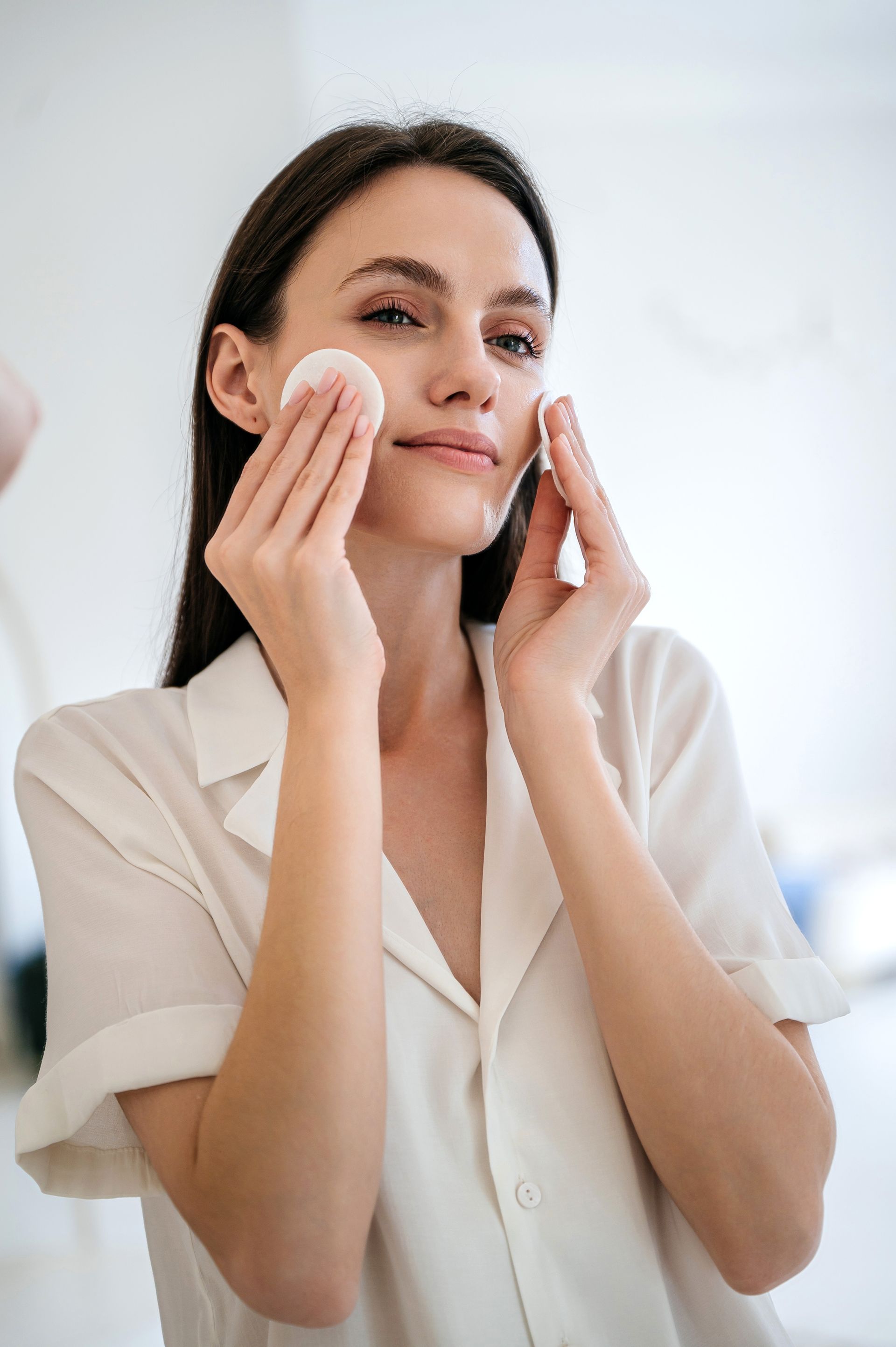 A woman is cleaning her face with a cotton pad in front of a mirror.