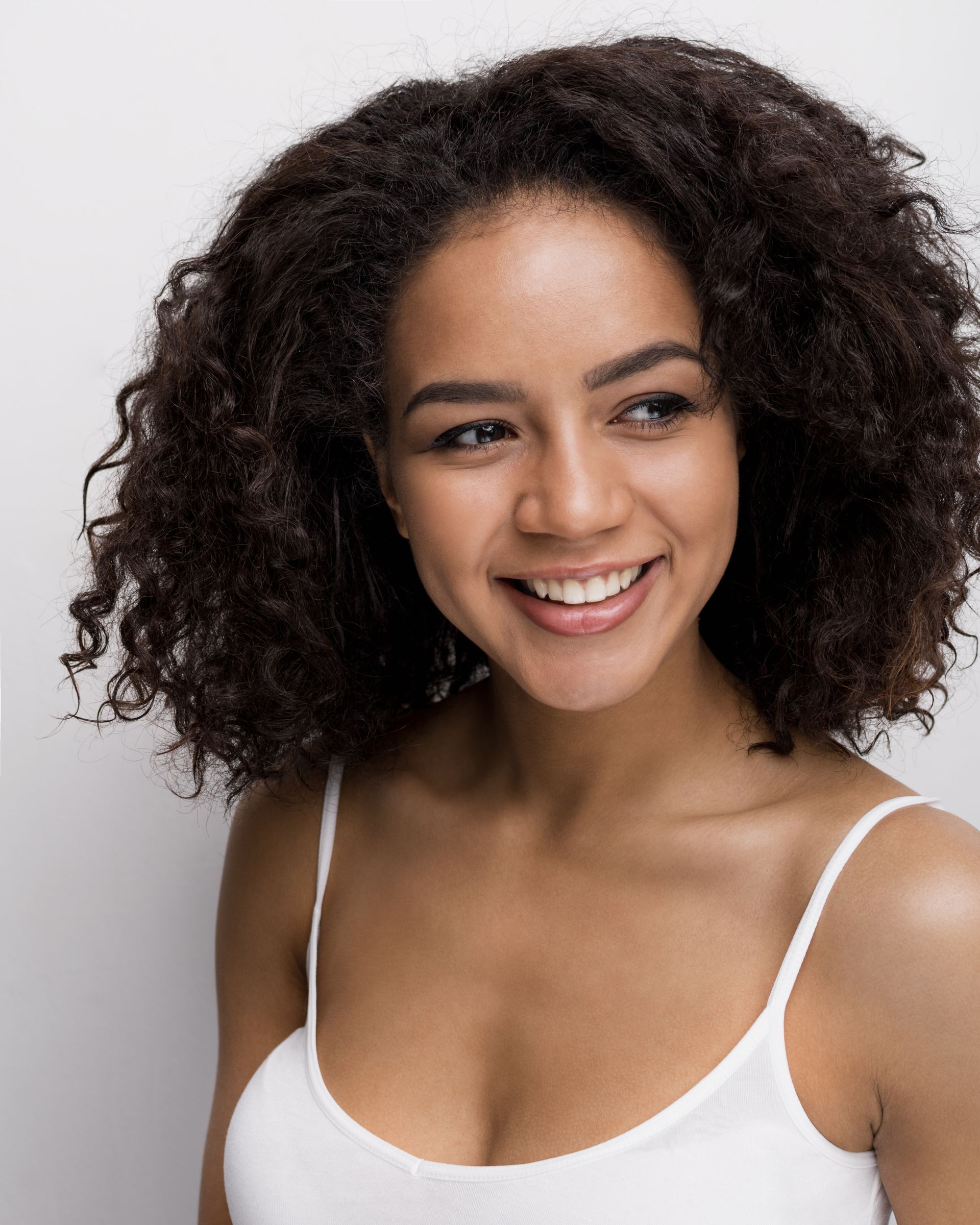 A woman with curly hair is smiling and wearing a white tank top.