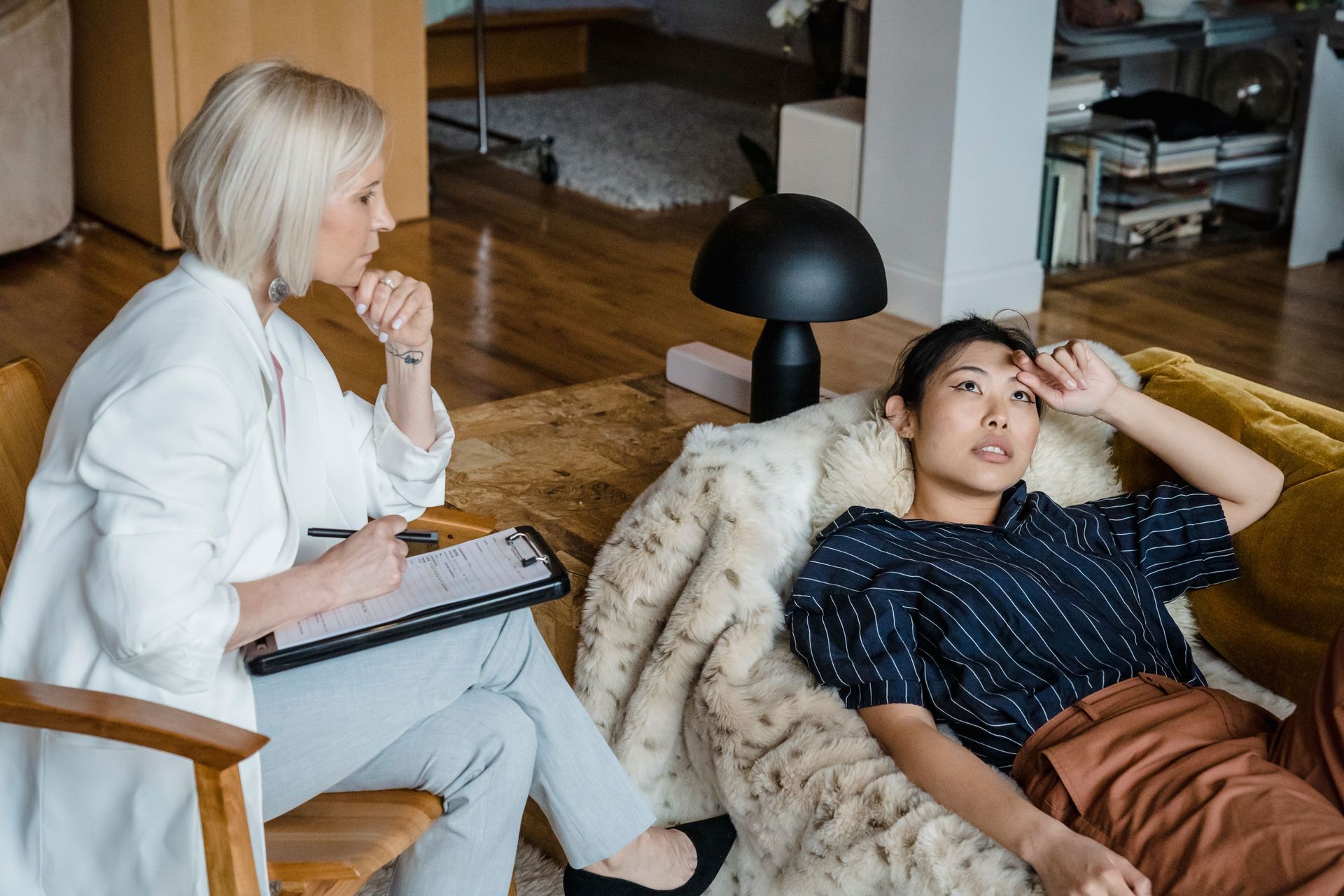 Two women in a living room, one taking notes while the other reclines on a chair and talks.