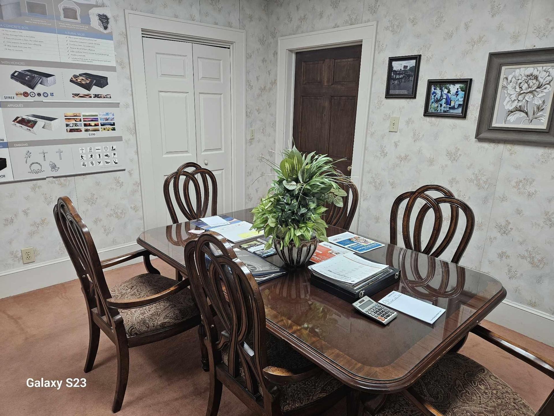 A meeting room with a dark wooden table, four chairs, a decorative plant, and framed pictures on patterned wallpaper.