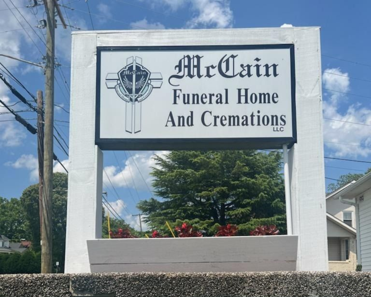 A white, rectangular sign for McCain Funeral Home and Cremations with a black cross logo against a blue sky.
