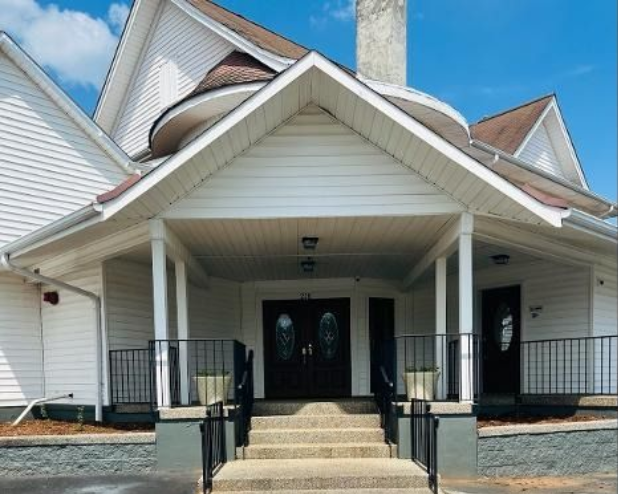 The front entrance of a white building with a covered porch, stone steps, and dark double doors under a gabled roof.