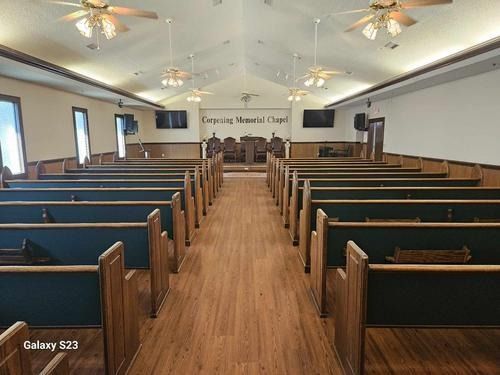 Empty chapel interior with rows of wooden pews facing a pulpit, ceiling fans, and wood-look flooring.
