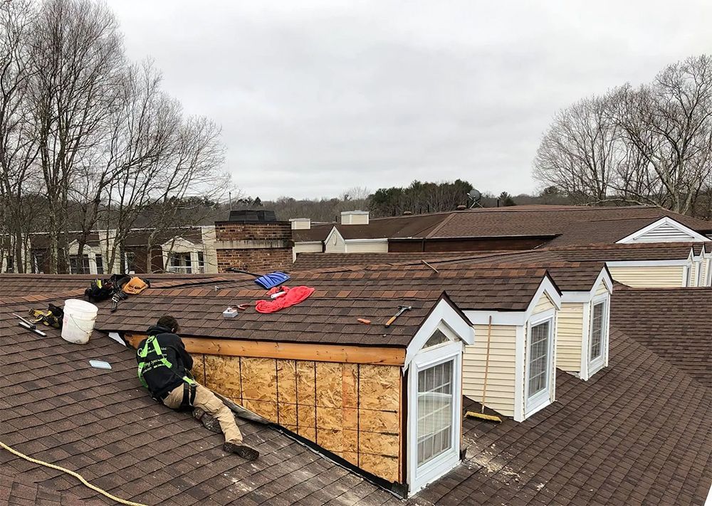 Roofers working on a brown shingle roof; cloudy day. One worker in harness and a partially finished wall.