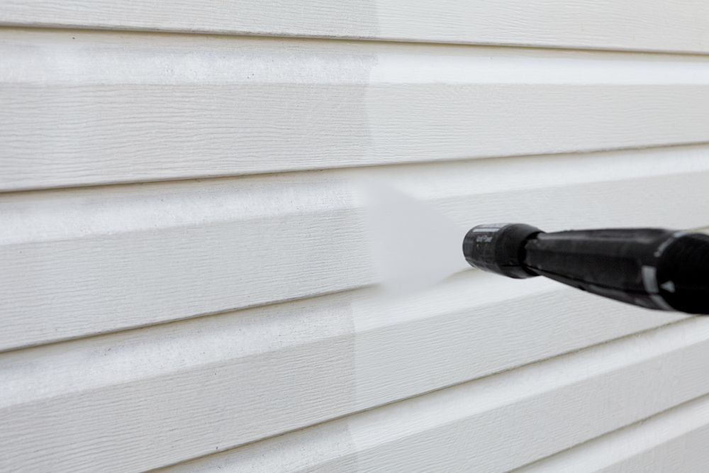 Pressure washer cleaning white siding; half is cleaned, half remains dirty.