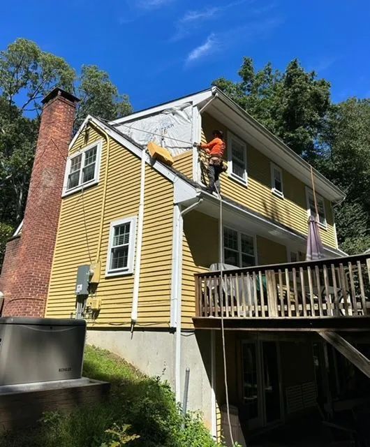 Yellow house with worker on roof repairing shingles, surrounded by green trees on a sunny day.