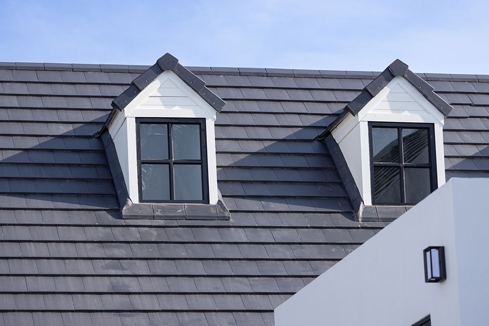 Two dormer windows on a gray tiled roof with white trim against a blue sky.