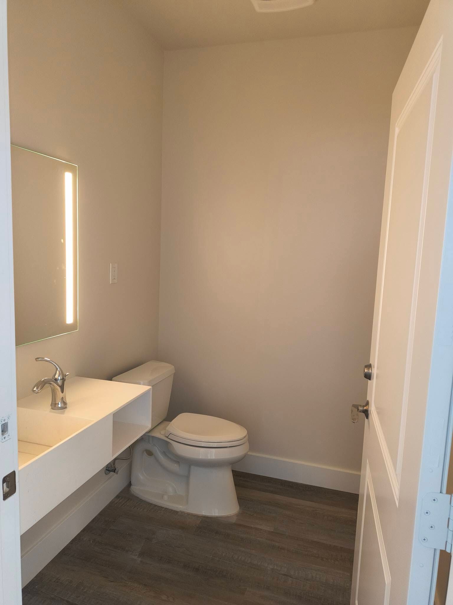 Small modern bathroom with floating white sink, illuminated mirror, toilet, and dark wood floor.