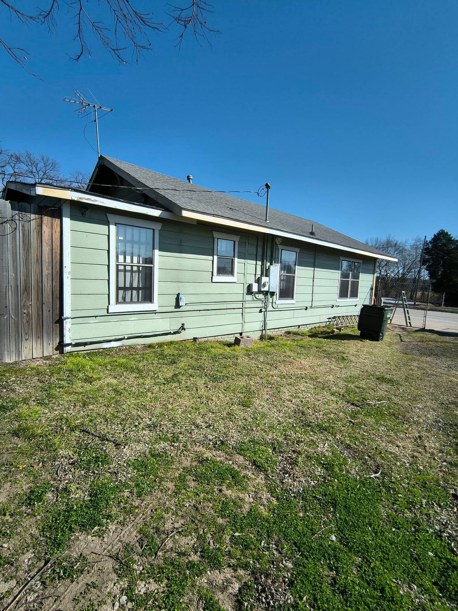 Light green single-story house with a sloped roof and fenced yard under a clear blue sky.