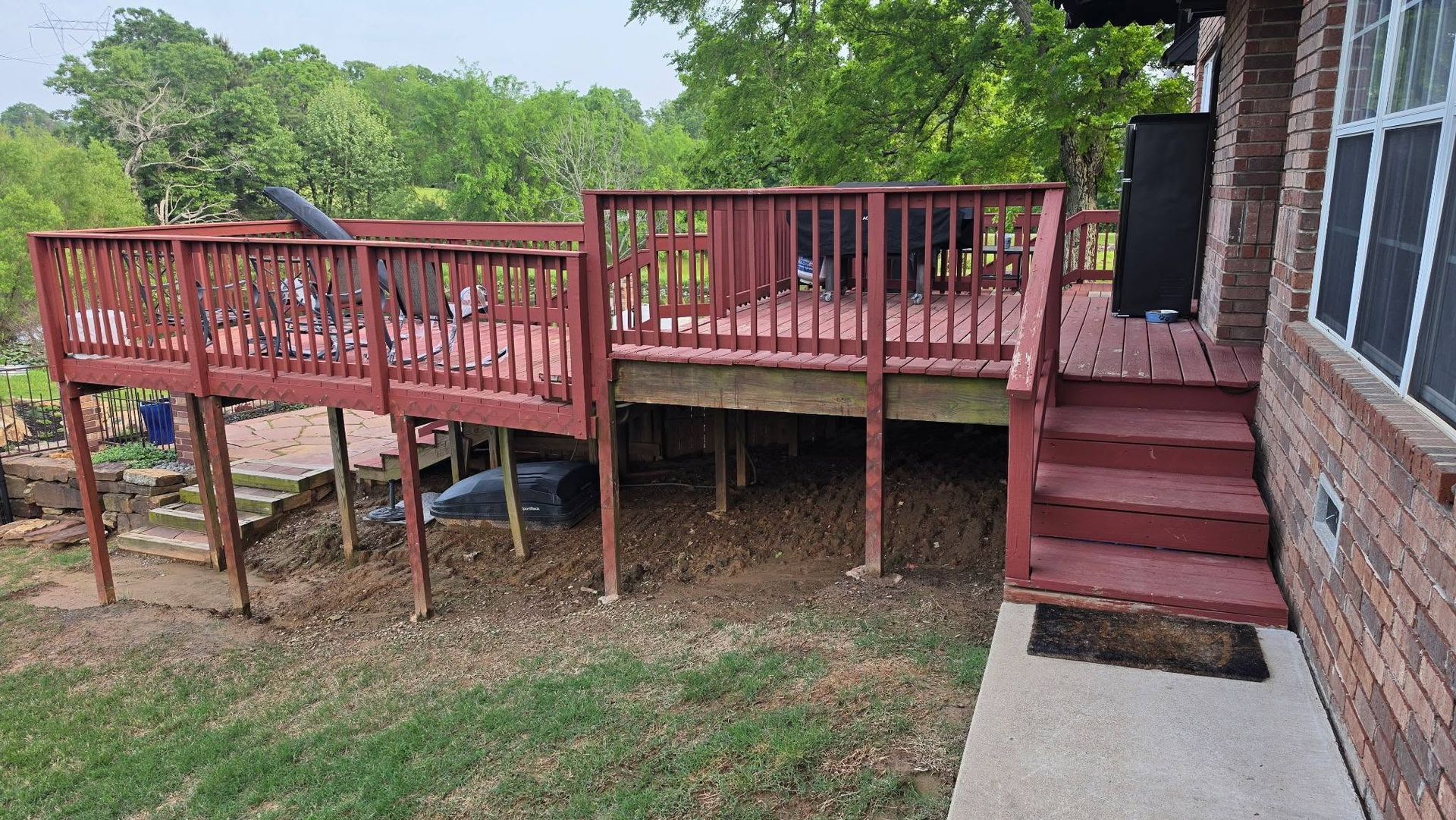 Red wooden deck with stairs beside a brick house, overlooking a grassy yard and trees