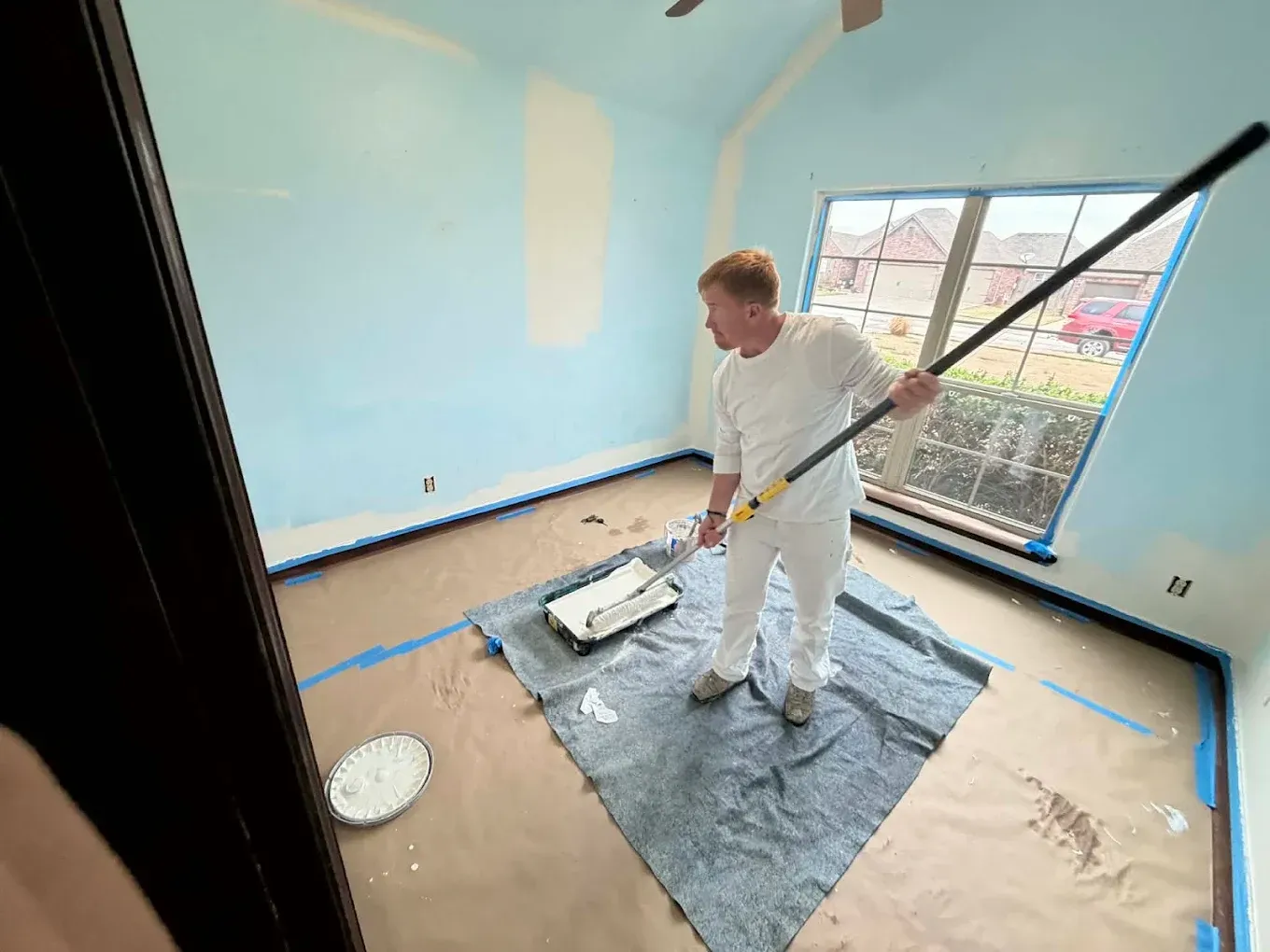 Person painting a light blue room with a roller, standing on a drop cloth near a window.