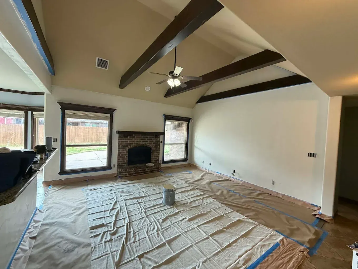 Living room under renovation with drop cloths, exposed ceiling beams, fireplace, and windows.