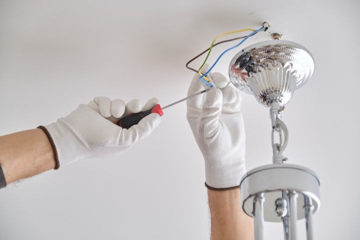Person installing a chrome light fixture on a white ceiling, wearing gloves and using a screwdriver.
