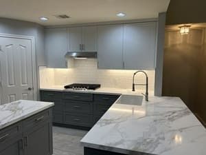 Modern kitchen with gray cabinets, marble countertops, stainless hood, and built-in cooktop.
