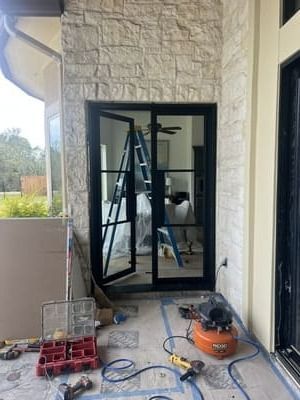 Contractor working on a black-framed glass door on a stone patio, with tools and materials nearby.