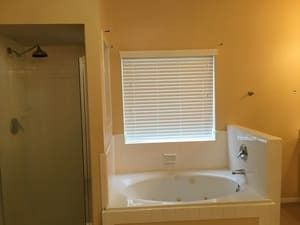Bathroom with a white bathtub, beige walls, a window blind, and a shower area on the left.