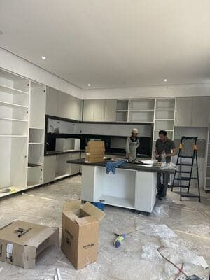 Renovated kitchen with white cabinets, island, and two workers amid tools and packing boxes