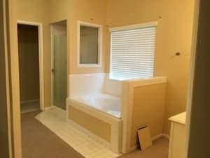 Bathroom with a white tub bench, window blinds, and beige walls with open doorways
