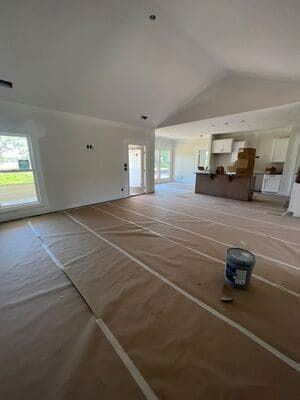 Empty room under renovation with protected floor, white walls, and a kitchen pass-through.