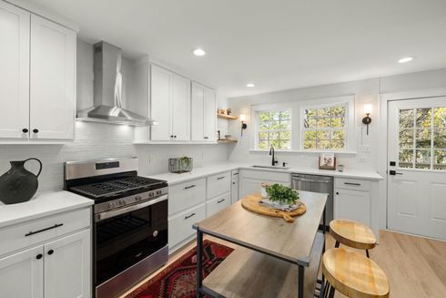 Bright modern white kitchen with black stove, wood island table, and stools by a window and back door
