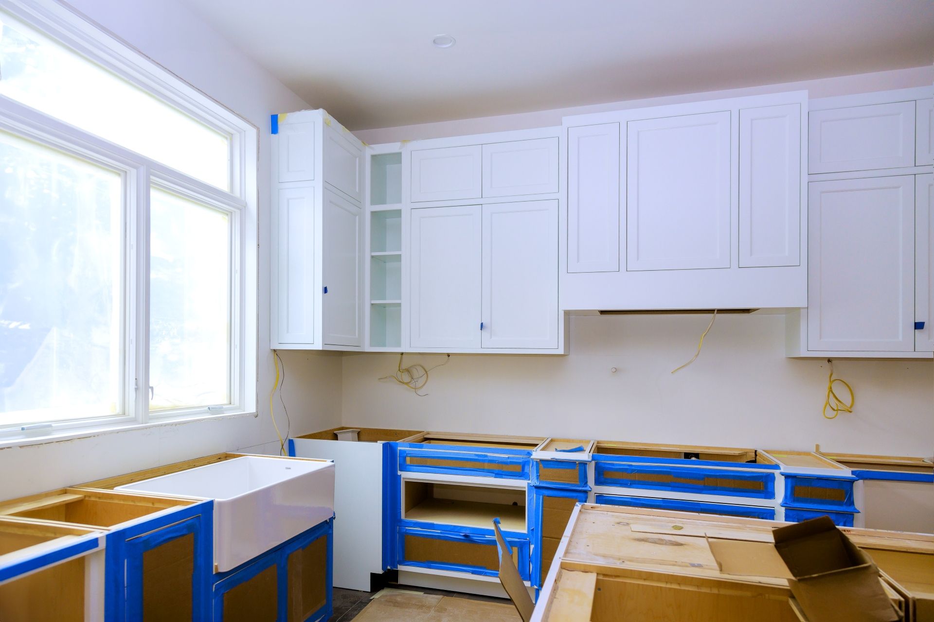 Bright kitchen under renovation with white cabinets, blue painter’s tape, and a large window.