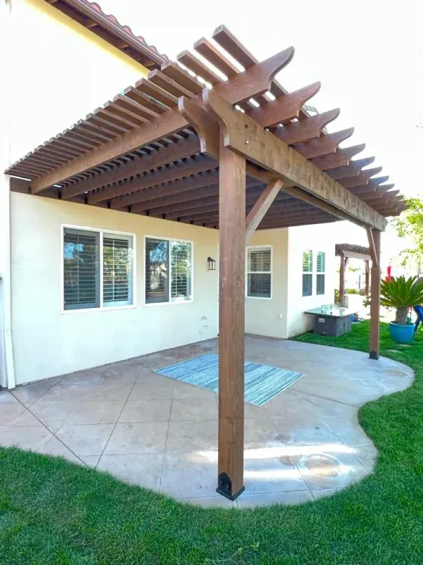 Wooden pergola covers a concrete patio next to a stucco house with a grassy yard.