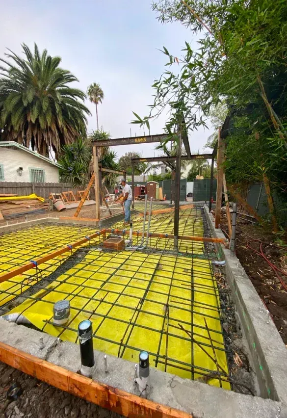 Construction site: Concrete foundation with yellow fabric, rebar, and wooden frames; a person works.