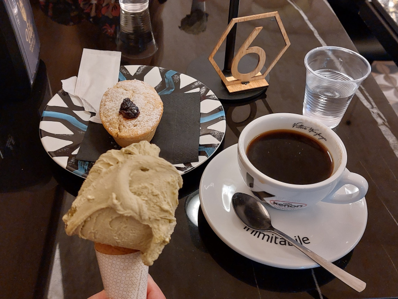 Su un tavolino di un bar, una mano regge un cono gelato al pistacchio accanto a un pasticcino, una tazza di caffè nero e un bicchiere d'acqua.