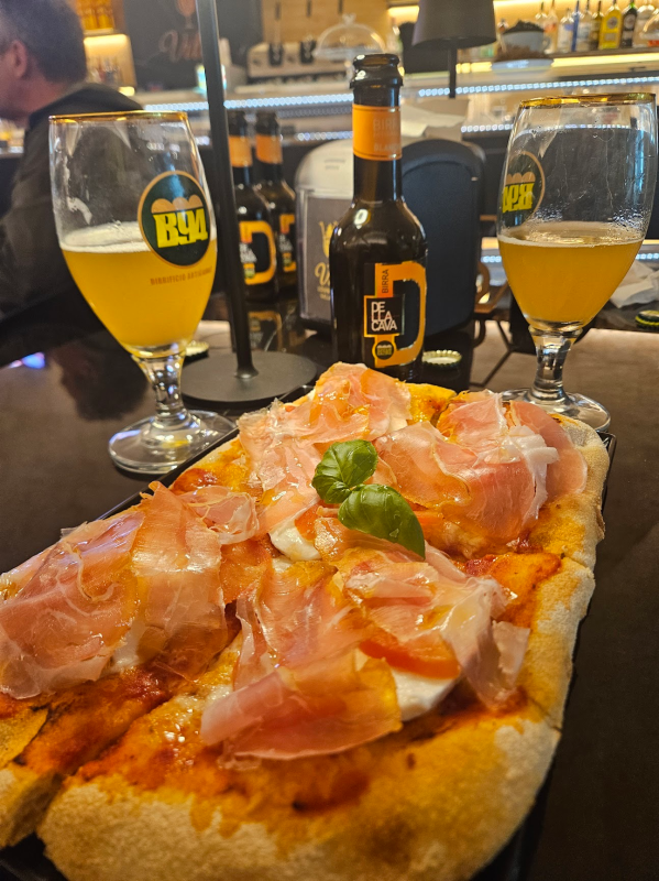 Una pizza rettangolare condita con prosciutto e basilico, servita con due bicchieri di birra e una bottiglia su un tavolino da bar.