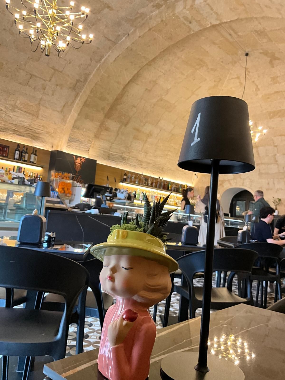Una statuetta con un vaso a forma di cappello giallo e una lampada da tavolo numerata 1 in un ristorante con soffitti a volta in pietra.